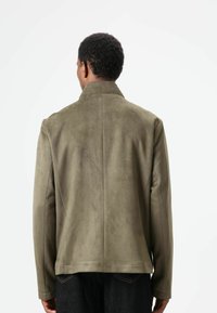 Olivgrüne Wildlederjacke mit hohem Kragen und tailliertem Schnitt. Glatte Textur, minimalistische Nahtdetails und Seitenpanele.