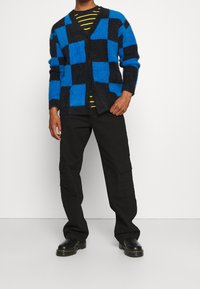 Sininen ja musta ruudullinen neule, jossa on pörröinen tekstuuri, napilla edessä, yhdistetty mustiin cargo-housuihin ja mustiin saappaisiin. Raidallinen paita alla.