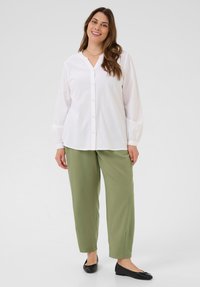 Femme souriante aux longs cheveux bruns portant une blouse blanche à boutons, un pantalon vert olive, des ballerines noires et un collier en or, se tenant devant un fond blanc.