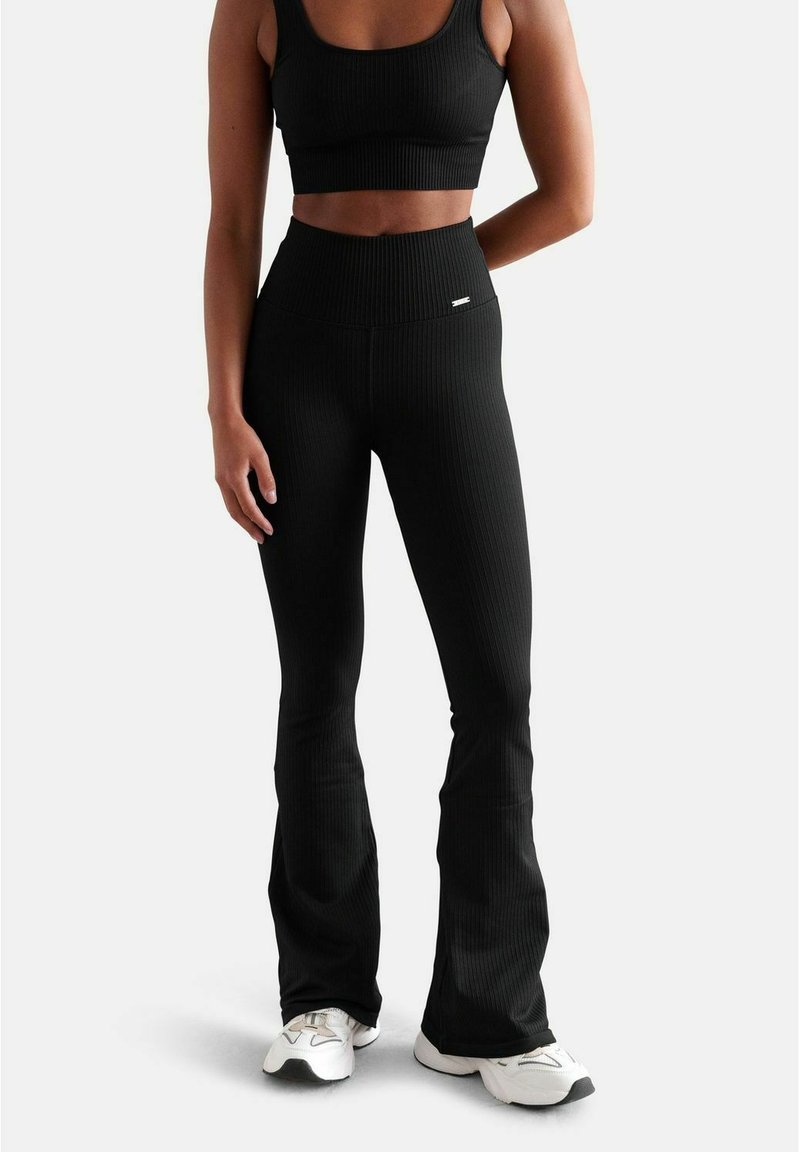 Schwarzes Rippensportoberteil und passende ausgestellte Leggings, die ein enganliegendes Design, hohe Taille und nahtlose Akzente aufweisen. Getragen mit weißen Sportschuhen.