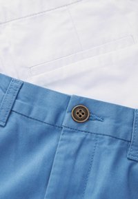 Vêtement en coton bleu avec une fermeture à bouton, présentant une texture lisse et un tissu blanc contrastant au dos, avec un détail de poche arrière.