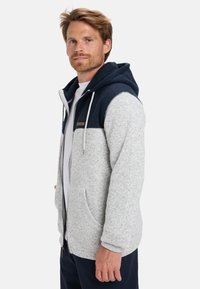 Sudadera con cremallera en gris y azul marino de acabado texturizado, con dos bolsillos delanteros, capucha con cordón y un detalle de piel sintética.