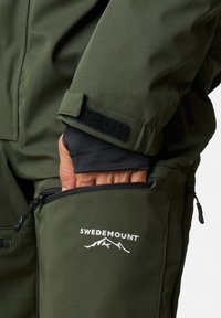 Zielona kurtka na zewnątrz z zamkiem błyskawicznym, rzepem przy mankietach i logo SWEDEMOUNT. Materiał wydaje się być trwały o gładkiej teksturze.