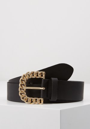 Ceinture en cuir noir avec une grande boucle en forme de maillon de chaîne dorée, enroulée sur une surface blanche sur un fond clair uni.