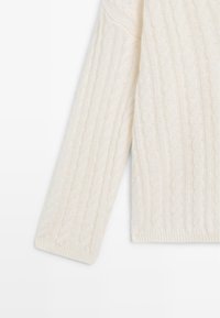 Maglione a treccia in crema, caratterizzato da motivi testurizzati e polsini a coste. Il tessuto appare morbido e leggermente strutturato, con un design accogliente.