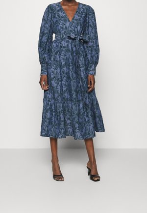 Femme portant une robe midi bleue à fleurs avec de longues manches bouffantes, un col en V, une taille ceinturée et des sandales à talons hauts noires ouvertes à l’avant.