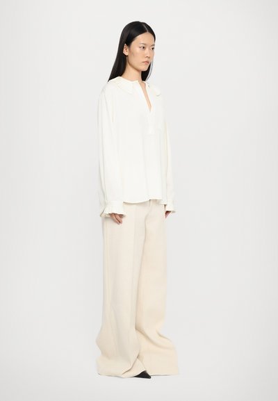 Cremfarbige Bluse mit plissiertem Kragen und weiten Ärmeln, kombiniert mit lässigen, hellen Hosen. Texturiertes Material, minimalistisches Design und entspannter Schnitt.