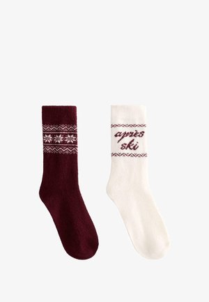 Pár vlnených ponožiek, jedna tmavočervená s bielym vzorom vločiek, druhá biela s červeným vzorom a textom "après ski", zimný štýl.