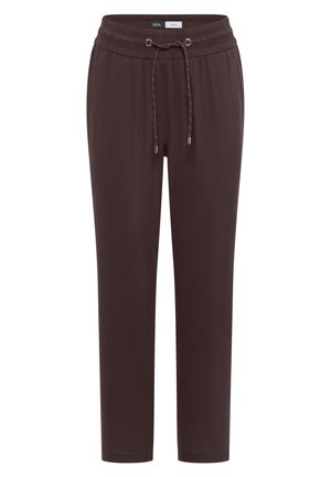 Frau lächelt, trägt ein blaues ärmelloses Top, braune Hose mit Kordelzug, hält eine große gewebte Tasche, steht vor einer beigen Wand.