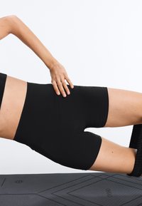 Person in schwarzen Shorts und Oberteil macht eine Seitenplanke auf einer Yogamatte, wobei eine Hand auf der Hüfte ruht und die Beine übereinandergestapelt sind.