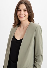 Lichtgroene blazer met een reverskraag, open voorkant en lange mouwen, gedragen over een zwarte top; gladde textuur en nauwsluitende pasvorm.