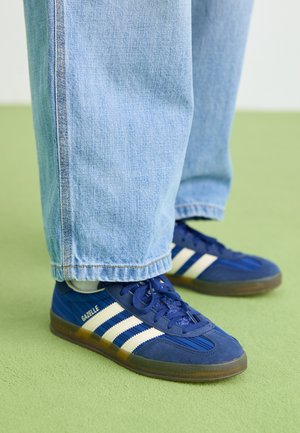 Sinised sametist tossud valgete triipudega ja tekstuursete mustritega, kantud helesiniste teksapükste ja rohelise tausta vastu.