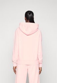 Hanorac roz supradimensionat dintr-un material moale, cu glugă pe spate, manșete și tiv ribbate. Designul are o estetică minimalistă.