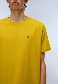 Keltainen puuvillapaita pyöreällä pääntiellä ja lyhyillä hihoilla. Rintakohdassa on pieni brodeerattu lippulogoe. Sujuva pinta.
