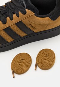 Bruna mockaskor med svarta ränder, runda svarta snören och en svart gummisula. Två bruna ovala skosnören visas nedanför.