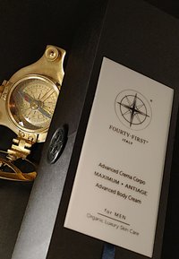 Gouden kompaswatch met zichtbare wijzerplaten en markeringen naast een witte doos met het label "EENENVEERTIGSTE" en "Geavanceerde Lichaamscrème" in zwarte tekst.