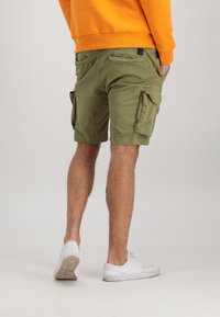 Personne portant un short cargo vert olive, un sweat-shirt orange et des baskets blanches, debout les mains dans les poches devant un fond uni.