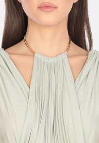 Collana choker in tonalità oro con design lucido e arrotondato. Tessuto plisettato verde chiaro pende sotto, creando un aspetto morbido e testurizzato.