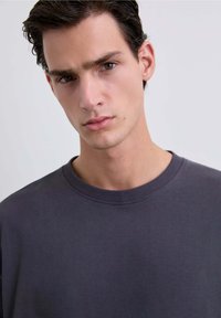 Sudadera gris oscuro con cuello redondo y detalles de puños acanalados. La tela parece suave y tiene una textura de peso medio.