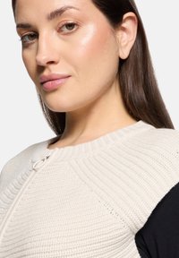 Pull en tricot avec un devant texturé crème et des manches noires unies. Il présente un col rond et un détail de fermeture à boutons sur le côté.