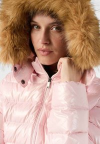 Mujer con una chaqueta acolchada de color rosa claro y una gran capucha forrada de piel marrón, sosteniendo la cremallera cerca de su mentón y mirando hacia adelante.