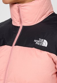 Ljusrosa jacka med svart övre panel. Har hög krage och dragkedja. Logotyp i vitt på vänster bröst. Texturerat tyg.