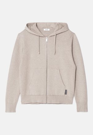 Mies, jolla on päällään beige vetoketjullinen huppari, valkoinen t-paita, tummanharmaat nyörillä varustetut housut, valkoiset lenkkarit ja musta olkalaukku.