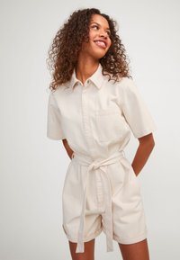 Mono de algodón beige con mangas cortas, un bolsillo, cintura ajustada y un cinturón atado. Los pantalones cortos tienen dobladillos enrollados para un detalle adicional.