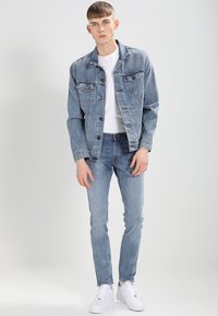 Hellblaues Jeansjacke mit zwei Brusttaschen und Knopfverschluss, getragen über einem weißen T-Shirt und kombiniert mit hellblauen Skinny-Jeans und weißen Sneakern.