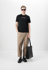 Black Calvin Klein t-shirt with red logo, khaki wide-leg pants, black sandals, and a black leather tote bag. Smooth fabric and minimalistic design.