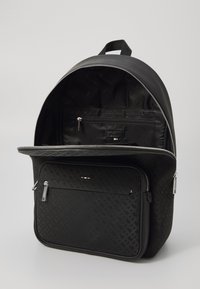 Sac à dos noir avec un motif texturé, deux zips, et un intérieur spacieux comportant une poche et une étiquette. Fabriqué en matériau synthétique.
