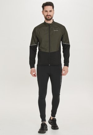 Schwarze und olivgrüne Sportjacke mit Reißverschluss vorne, kontrastierenden Ärmeln und reflektierenden Akzenten, kombiniert mit schwarzen, enganliegenden Hosen und Turnschuhen.