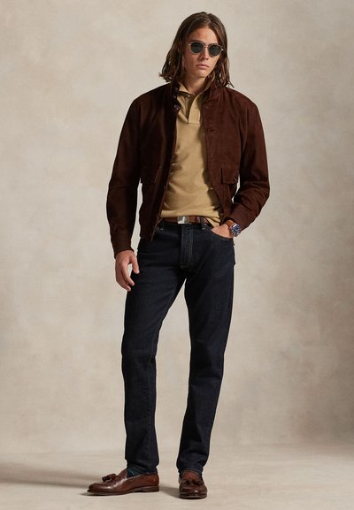 Moški, ki nosi temnordeč usnjen suknjič, bež polo majico, temne kavbojke, rjave loaferje s cofki, okrogle sončna očala in ročno uro, stoji na nevtralnem ozadju.