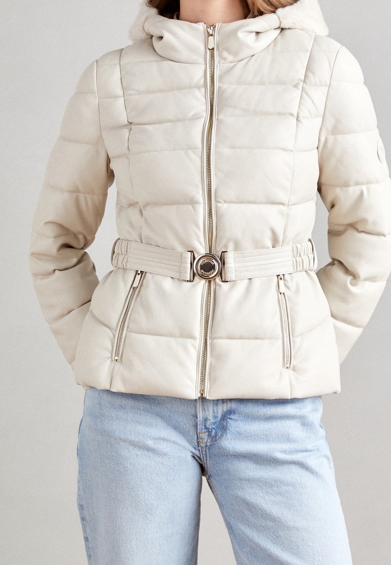 Beige quiltet jakke med hætte, der har en bæltet talje med en cirkulær spænde og for-zip lommer. Glat, blank overflade.