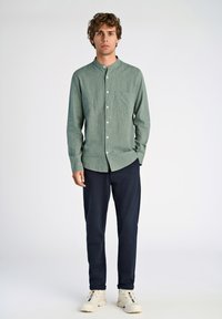 Camisa abotonada verde claro con cuello alto, bolsillo en el pecho, acompañada de pantalones azul marino oscuros y zapatillas blancas. Detalle de puños ajustables.