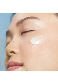 Crème hydratante appliquée sur la joue ; texture lisse et crémeuse ; fini léger et réfléchissant sur un teint bronzé et uniforme.