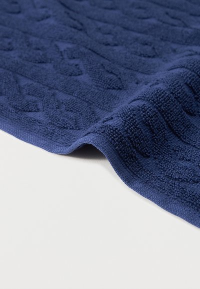 Ύφασμα πλεκτό σε ναυτικό μπλε με υφή και ανάγλυφο γεωμετρικό σχέδιο καλωδίου, μερικώς διπλωμένο σε λευκή επιφάνεια.