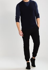 Marinblå v-ringad tröja över en mörk långärmad skjorta, i kombination med svarta joggarbyxor och svarta hög-top sneakers. Enkel, avslappnad outfit.