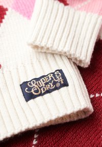 Guanti a coste di colore crema con un'etichetta nera e dorata "Superdry" su uno sfondo di tessuto a motivo rosso e rosa.