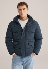 Ung man som bär en marinblå pufferjacka med huva, vit skjorta och ljusblå jeans, står med händerna i fickorna mot en enkel bakgrund.