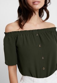 Paita olkapäältä avattuna, lyhyet hihat tummanvihreästä kankaasta. Siinä on tekstuuri pinta ja keskellä napit kiinnitystä varten.