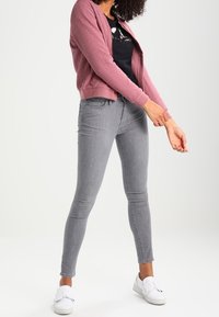 Chaqueta rosa claro con textura, cerrada con cremallera, sobre una camiseta gráfica negra, combinada con jeans gris claros de corte slim y zapatillas blancas sin cordones.