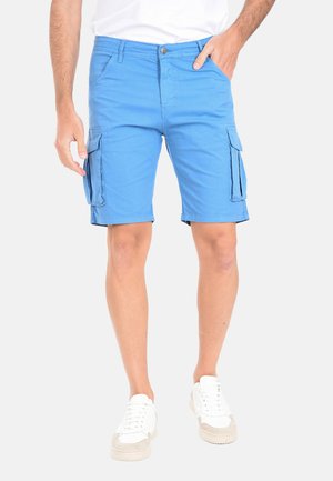 Uomo che indossa pantaloncini cargo azzurri chiari e sneaker bianche, con una mano in tasca, in piedi contro uno sfondo bianco.