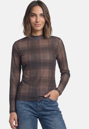 Femme aux cheveux bruns mi-longs portant un haut marron à carreaux à manches longues et un jean bleu, une main dans la poche.