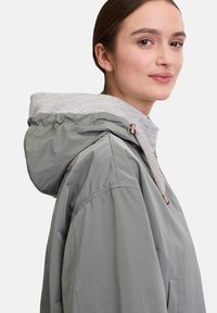 Lichtgrijze jas met capuchon, voorzien van een contrasterende grijze binnenvoering, voorzakken en een trekkoorddetail. Glad, waterafstotend materiaal.