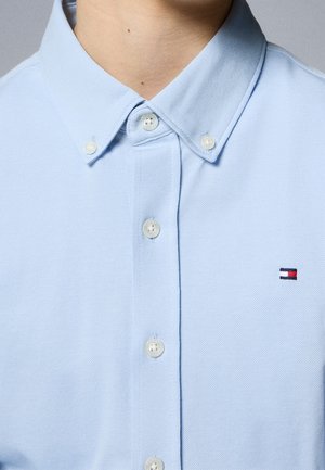 Jasnoniebieska koszula zapinana na guziki z kołnierzykiem na guziki, noszona przez osobę, z małym prostokątnym logo w kolorach czerwonym, białym i granatowym na piersi.