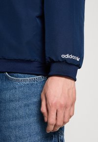Mörkblå Adidas-jacka med elastiska ärmslut, slät textur och vit logodetalj på ärmen. Bärs med denimjeans.