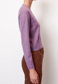 Lila, lätt cardigan med långa ärmar, v-ringning och knappar fram. Buren över bruna byxor, som visar en figurnära silhuett.