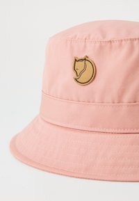 Pink bucket hat made of fabric with a textured finish. Features a circular, tan logo depicting a fox on the front. Wide brim design.