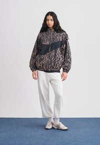 Jastučasta jakna s printom leoparda i crnim detaljima, u kombinaciji sa svijetlosivim trenirkama i neutralnim tenisicama, na jednostavnoj pozadini.